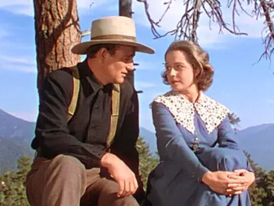 John Wayne and Betty Field sitting on a rock in a picturesque countryside setting in the 1941 film, The Shepherd of the Hills.