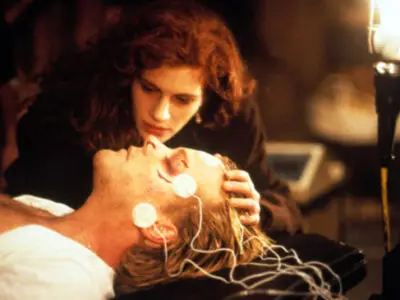 Julia Roberts nurses an unconscious Kiefer Sutherland, who lies on a hospital bed with monitors attached to his head and face, in the film, Flatliners.