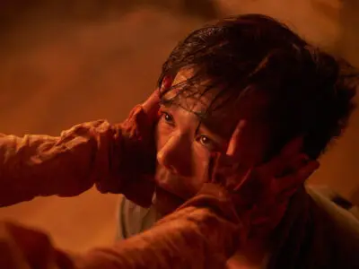 A man, lit in a red-orange fiery glow, looks in pain and fear as two hands grip his temples.