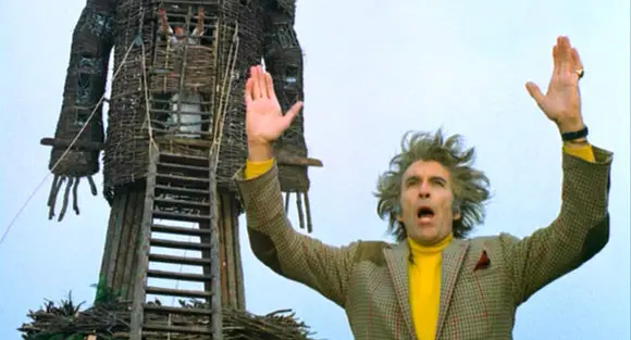 In front of a wicker man, Christopher Lee, his arms held high and his mouth wide open, as he speaks to his followers in the 1973 film The Wicker Man.