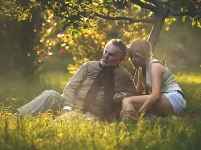 Ellie Bamber and Derek Jacobi sit on the ground in a summer garden surrounded by yellow flowers in the film, Moss and Freud.