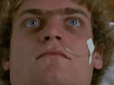 An image from the film Patrick which sees a man in a hospital bed, his eyes wide open, and a nasogastric tube inserted in his nose and secured by white tape on his left cheek.