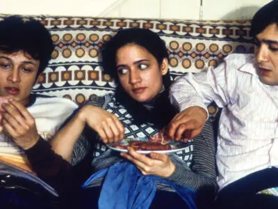 Jimi Mistry as Tariq "Tony" Khan in the 1999 film, East is East, sits on a 1970s couch alongside fellow actors Chris Bisson and Archie Panjabi.